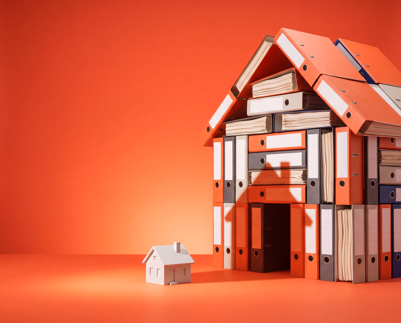Ein Haus, gebaut aus Aktenordnern — davor ein winziges Modellhaus, das einen langen Schatten wirft.